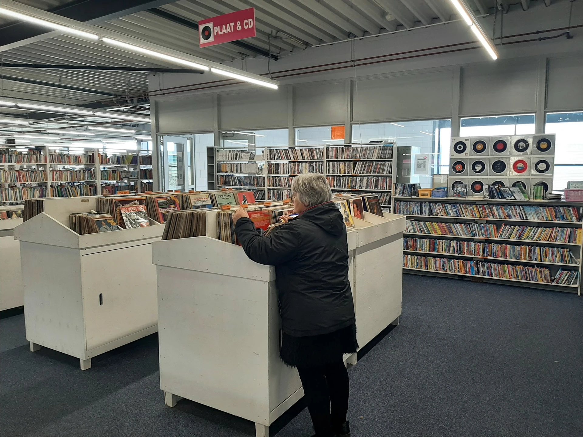 Vrouw zoekt tussen cd's in een kringloopwinkel met stellingen vol boeken en cd's op de achtergrond