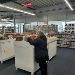 Vrouw zoekt tussen cd's in een kringloopwinkel met stellingen vol boeken en cd's op de achtergrond