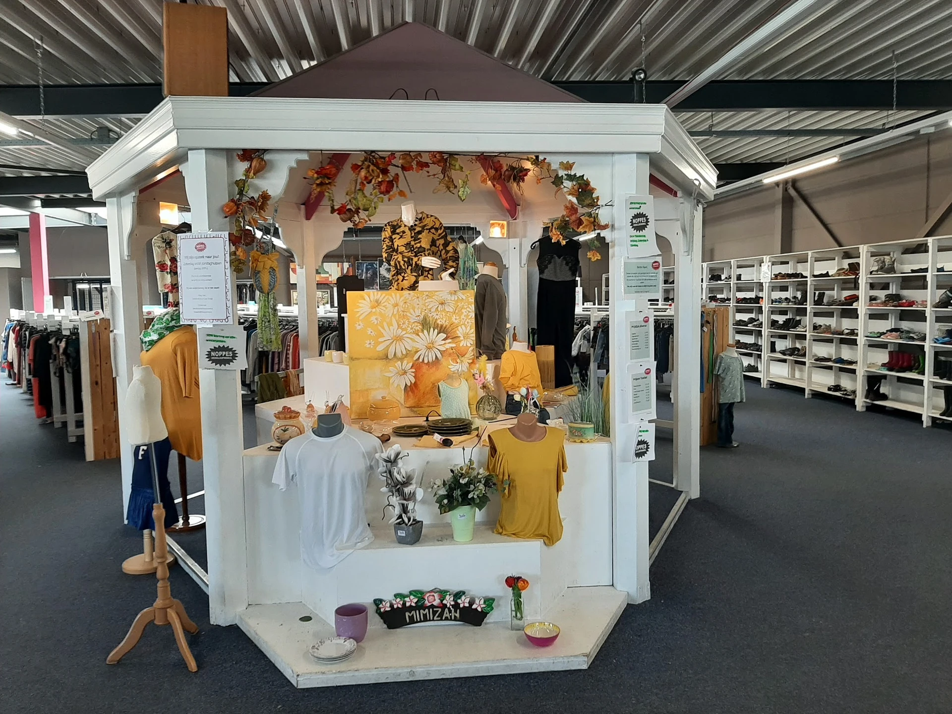 Uitgestalde kledingcollectie in een wit display met herfstdecoratie, bloemen en gele kledingstukken op mannequins