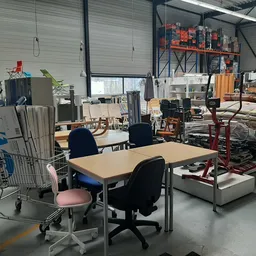 Werkruimte in kringloopwinkel met tafel, bureaustoelen, stellingen met spullen en diverse items