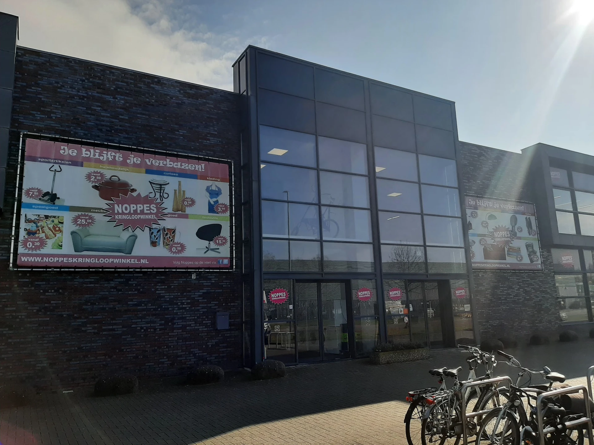 Voorgevel van kringloopwinkel met zwarte bakstenen muur, groot raam met rood logo, advertentiebord links en fietsen geparkeer