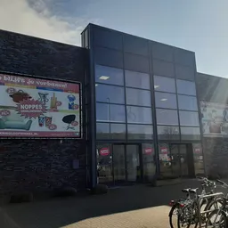 Voorgevel van kringloopwinkel met zwarte bakstenen muur, groot raam met rood logo, advertentiebord links en fietsen geparkeer