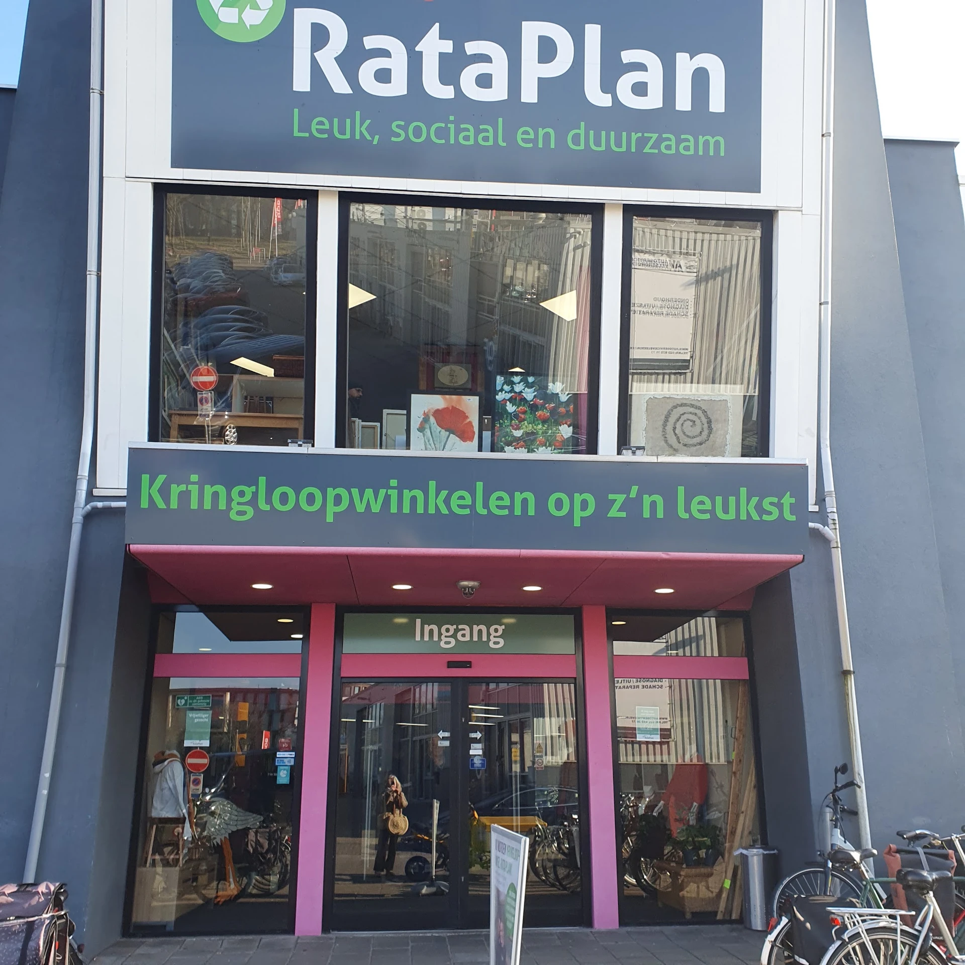 Gevel van kringloopwinkel met roze entree, groene tekst op witte bovengevels en etalages met diverse spullen