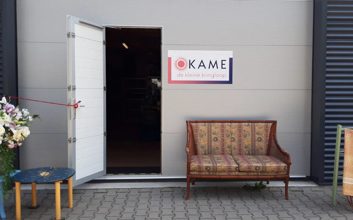Winkelentree met open deur, logo van Okame de kleine kringloop, en vintage meubels op het terras