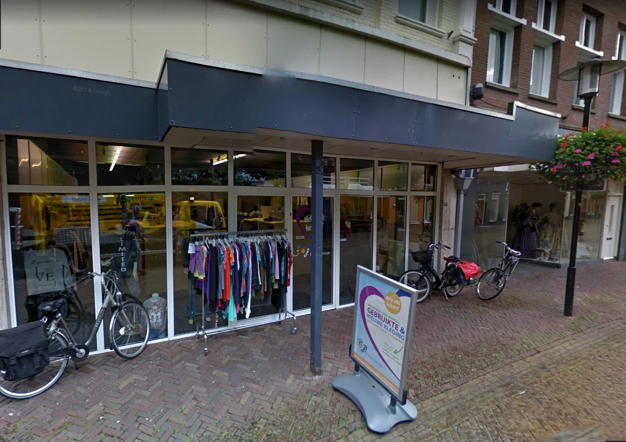 Voorgevel van kringloopwinkel met etalageruiten, tweedehands kleding aan rekken, fietsen buiten, donker afdak en straattegel
