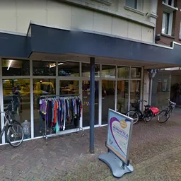 Voorgevel van kringloopwinkel met etalageruiten, tweedehands kleding aan rekken, fietsen buiten, donker afdak en straattegel