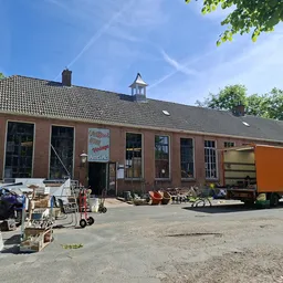 Voorkant van een bruinen bakstenen kringloopwinkel met dakkapel, omgeven door uitgestalde spullen op het terrein en een oranj