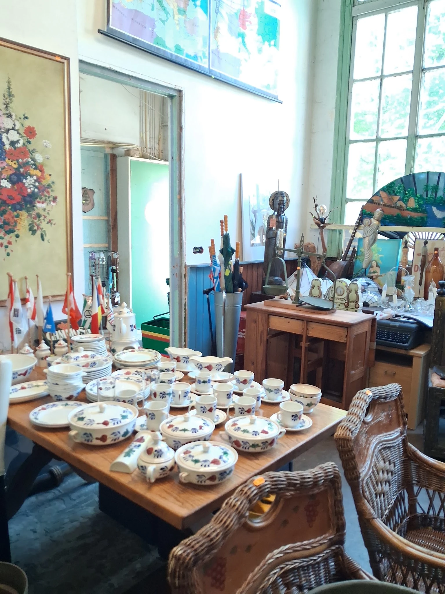 Uitgestalde servies met bloemmotieven op houten tafel in kringloopwinkel, met diverse antieke voorwerpen en decoraties op ach