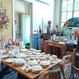 Uitgestalde servies met bloemmotieven op houten tafel in kringloopwinkel, met diverse antieke voorwerpen en decoraties op ach