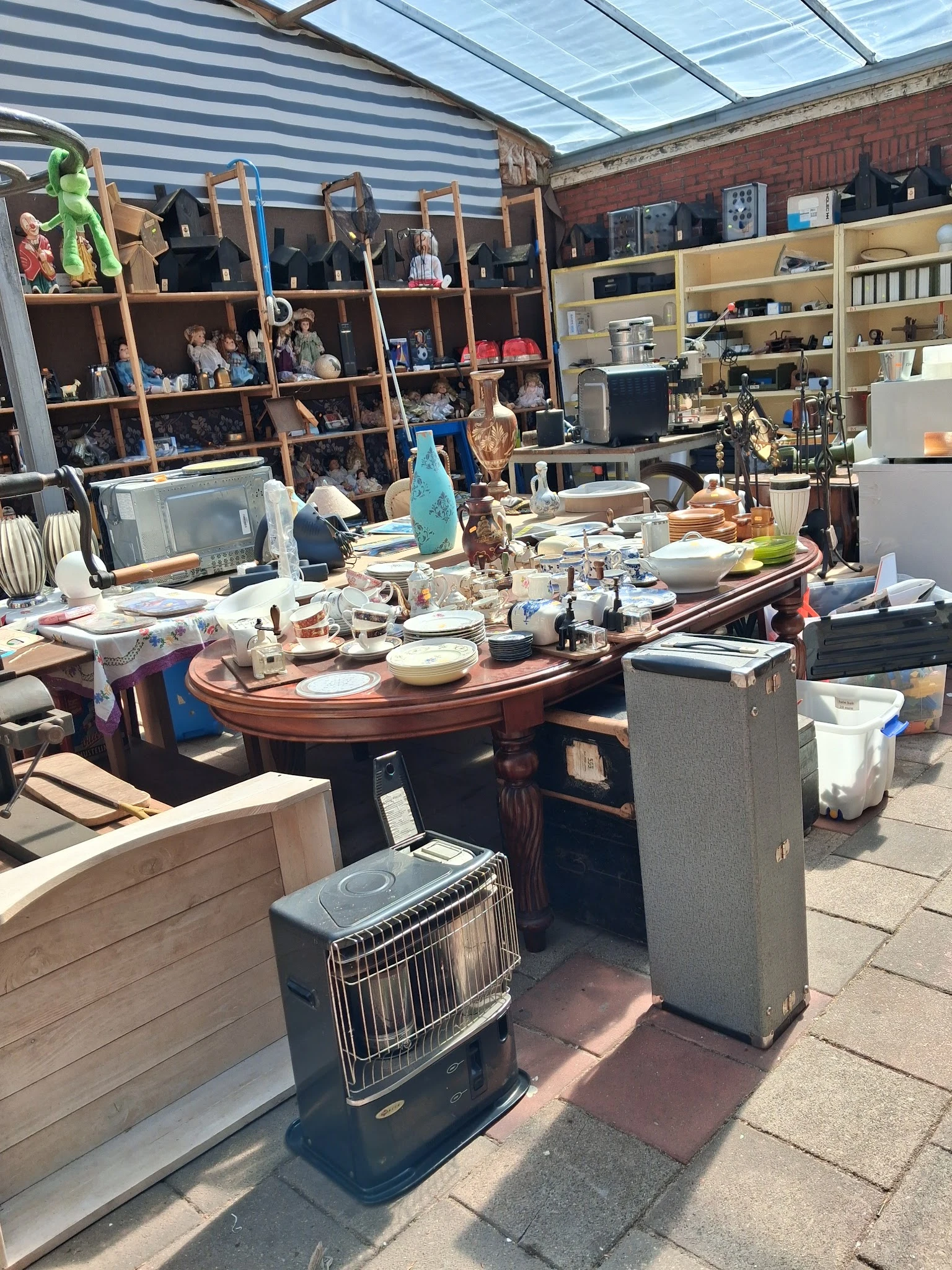 Binnenaanzicht van kringloopwinkel met houten bureaus, tafels, stellingen vol spullen en diverse meubels en huishoudartikelen