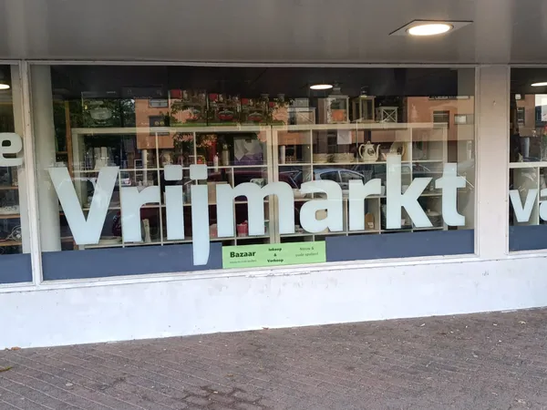 Winkelruit met witte letters 'Vrijmarkt' op donkerblauwe ondergrond, met zicht op winkelinterieur met stellingen
