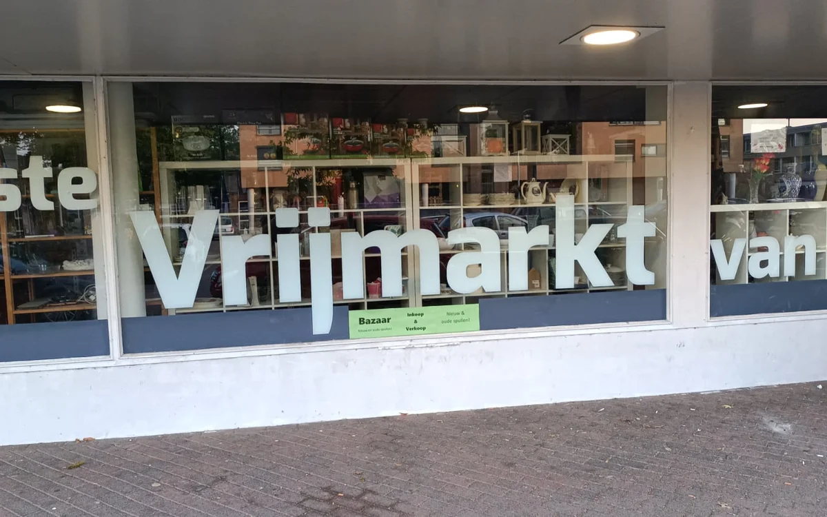 Winkelruit met witte letters 'Vrijmarkt' op donkerblauwe ondergrond, met zicht op winkelinterieur met stellingen