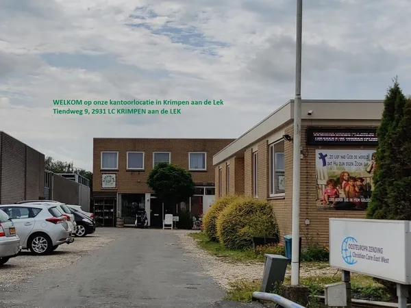 Voorkant van kringloopwinkel met bruine bakstenen gevel, parkeerplaats met auto's en groene beplanting