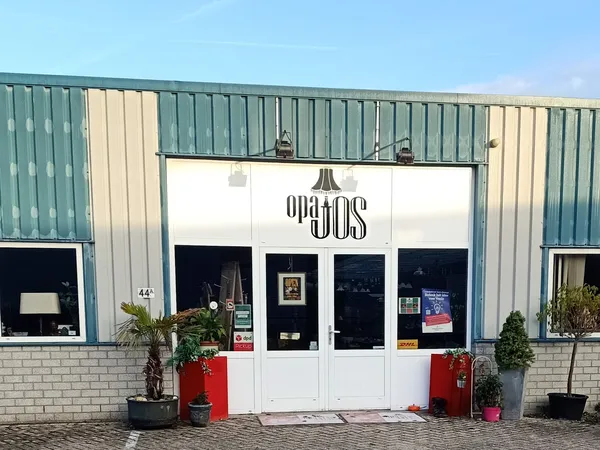 Witte winkelgevel met donkere ramen en 'OPA JOS' letters, planten in rode bakken aan weerszijden van de ingang