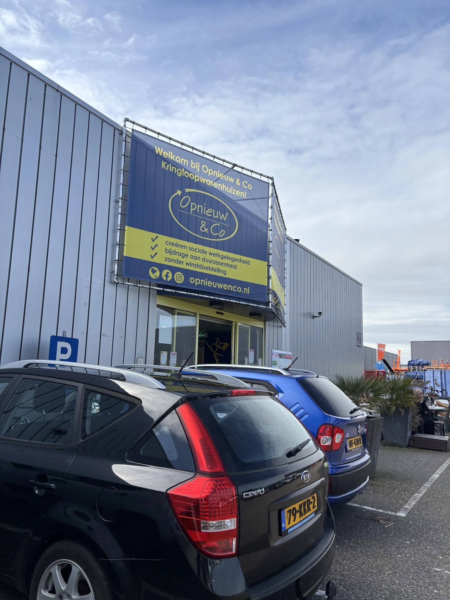Gevel van kringloopwinkel Opnieuw & Co met blauwe zijluifel, winkelnaam en logo zichtbaar, parkeerplaats met auto's ervoor