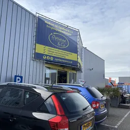 Gevel van kringloopwinkel Opnieuw & Co met blauwe zijluifel, winkelnaam en logo zichtbaar, parkeerplaats met auto's ervoor