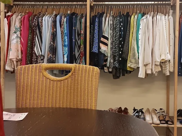 Twee kledingrekken met uitgestalde tweedehands kleding in verschillende kleuren en een rieten mand op een tafel.