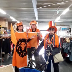 Drie vrijwilligers in oranje kleding en accessories poseren in de winkel