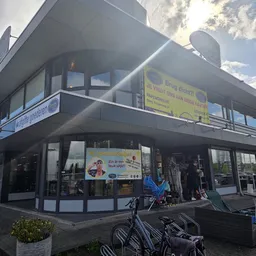 Hoekpand van kringloopwinkel met glazen pui, etalageruiten en gele borden boven met winkelinformatie