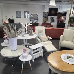 Interieur van kringloopwinkel met witte en houten ronde tafels, crèmekleurige stoelen, rode bank op achtergrond en diverse me