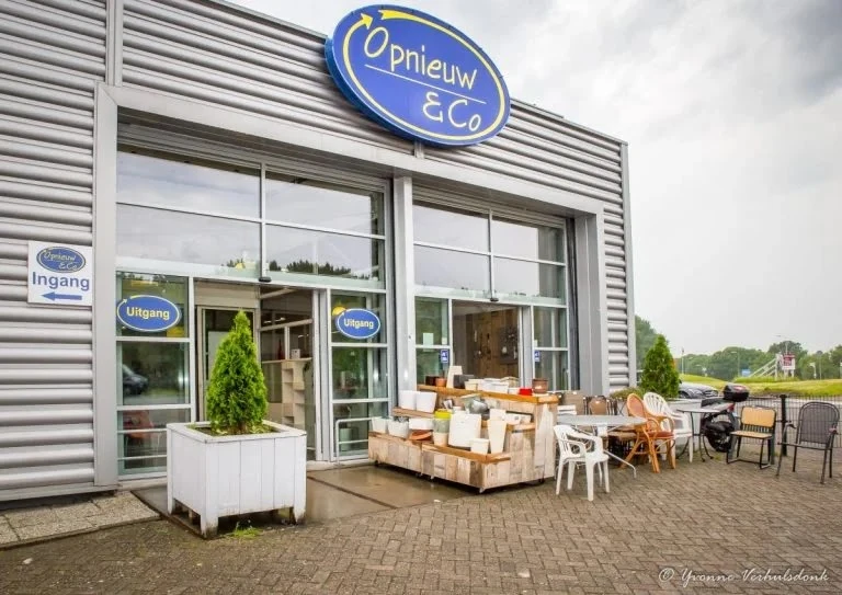 Voorkant kringloopwinkel met blauwe ovale logo 'Opnieuw & Co', glazen deuren, uitgestalde spullen op terras