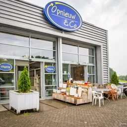 Voorkant kringloopwinkel met blauwe ovale logo 'Opnieuw & Co', glazen deuren, uitgestalde spullen op terras
