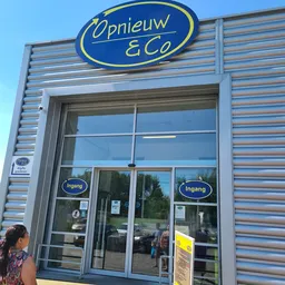 Gevel van kringloopwinkel met blauwe kunststof golfplaten, twee glazen deuren met 'Ingang'-teksten en blauwe ovaal logo boven