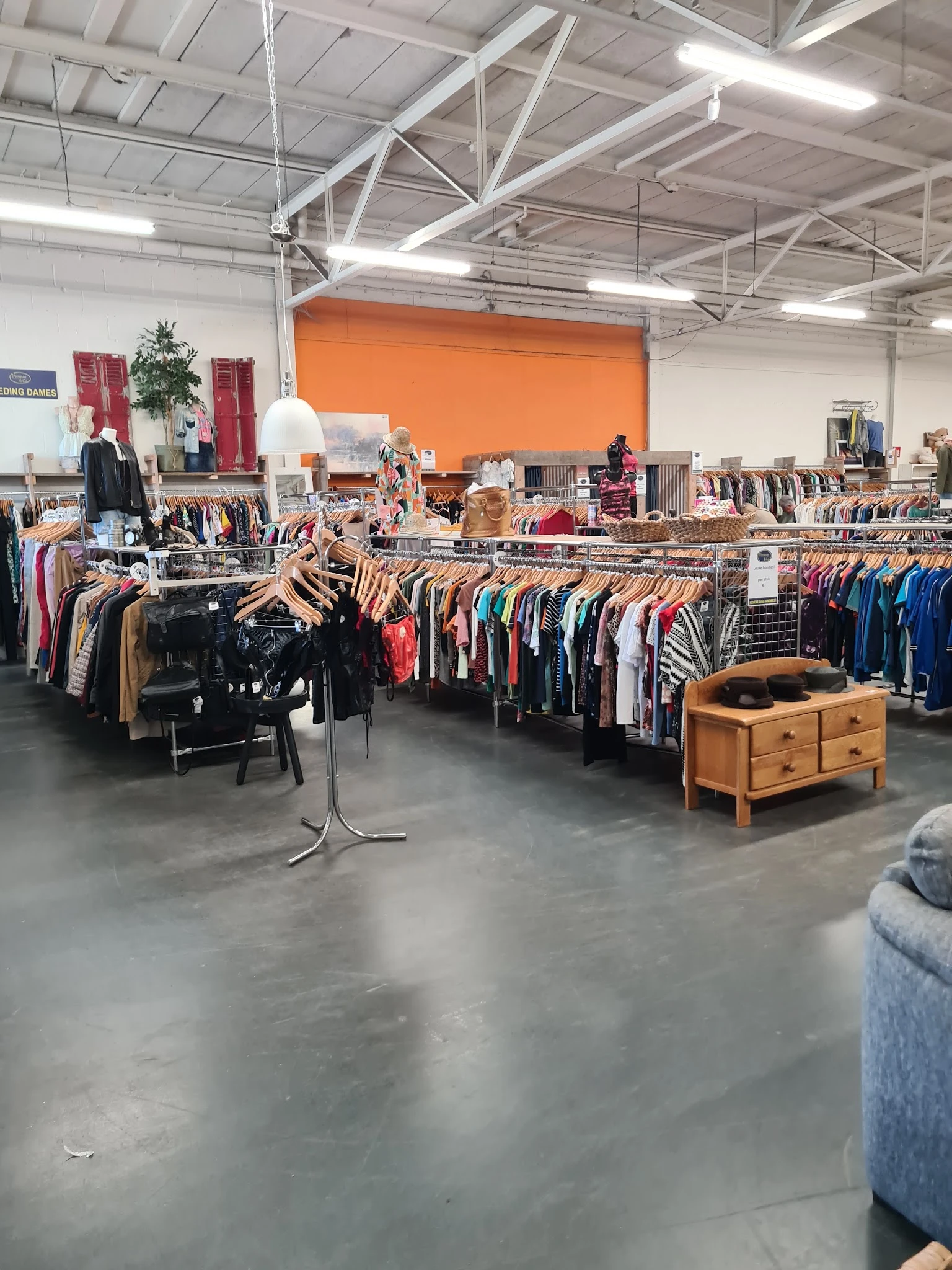 Winkelvloer met kledingrekken vol tweedehands kleding, houten meubel op voorgrond, oranje muur op achtergrond