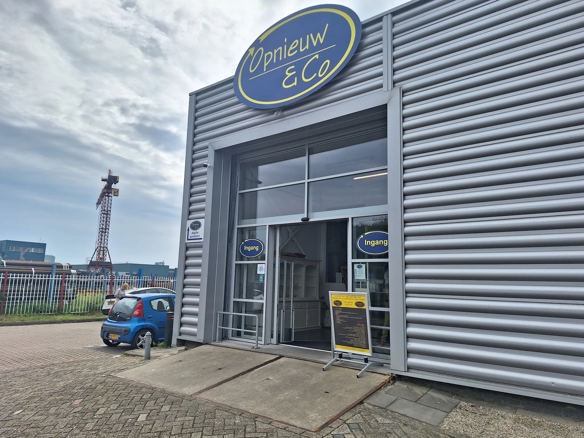 Grijze gevel van kringloopwinkel Opnieuw & Co met blauwe ovaal logo, ingang met glazen deuren en ramen, kleine blauwe scooter