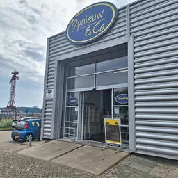 Grijze gevel van kringloopwinkel Opnieuw & Co met blauwe ovaal logo, ingang met glazen deuren en ramen, kleine blauwe scooter
