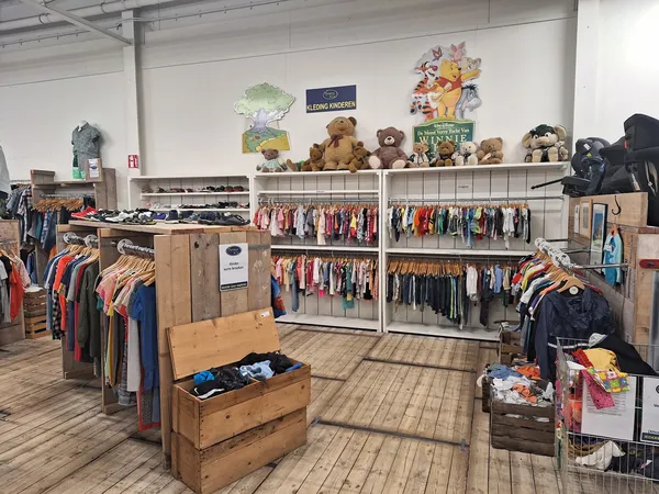 Ruim assortiment tweedehands kleding op rekken en hangers, met knuffels en speelgoed op hoge stellingen