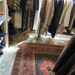 Winkelvloer met Perzisch tapijt, kledingrekken met vintage jassen en broeken, zonnig interieur