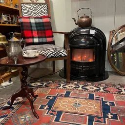 Tweedehands interieurstukken: houten tafel, rood-zwart geruit kussen, zwarte elektrische kachel, ornamentaal tapijt en spiege