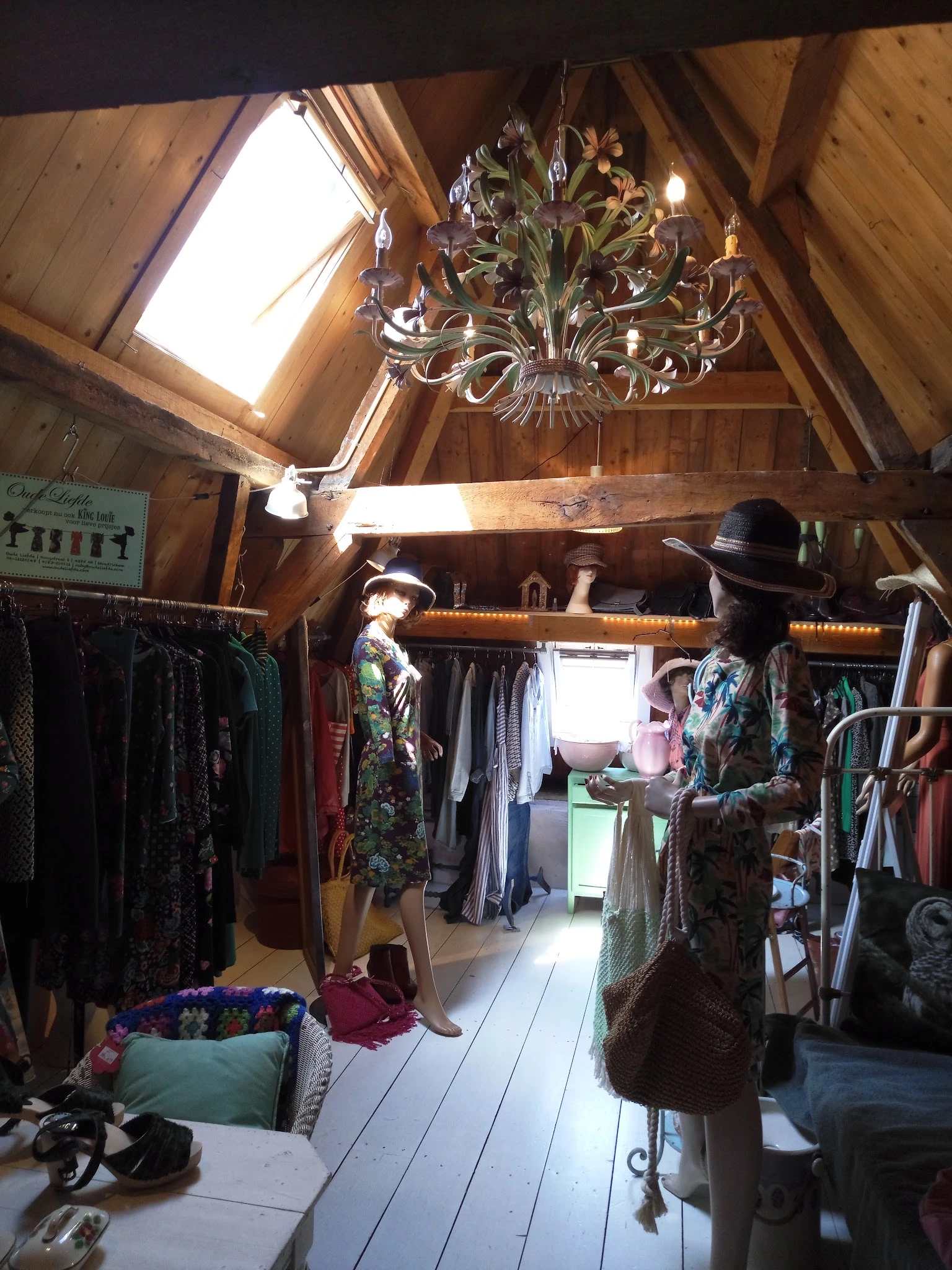 Tweedehands kledingwinkel met houten zolderruimte, kledingrekken, een kroonluchter en een mannequin met hoed