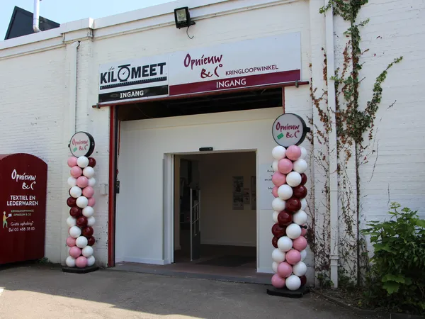 Ingang van kringloopwinkel met wit gevel, rode accenten en roze-witte ballonnen aan beide zijden van de deur