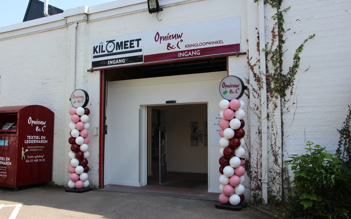 Ingang van kringloopwinkel met wit gevel, rode accenten en roze-witte ballonnen aan beide zijden van de deur