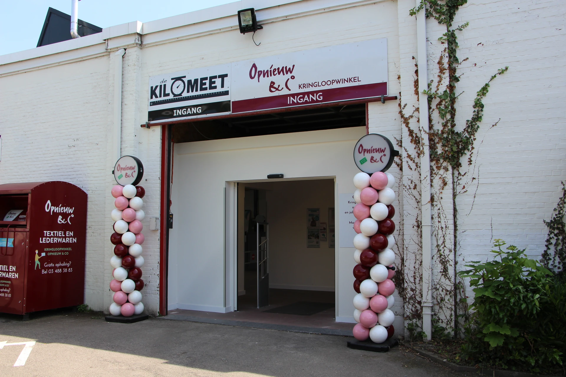 Ingang van kringloopwinkel met wit gevel, rode accenten en roze-witte ballonnen aan beide zijden van de deur