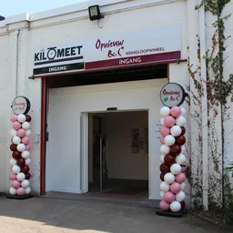 Ingang van kringloopwinkel met wit gevel, rode accenten en roze-witte ballonnen aan beide zijden van de deur