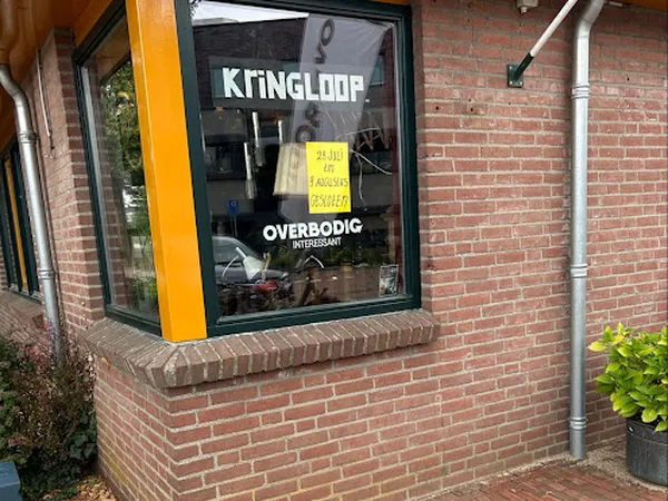 Winkelraam met KRINGLOOP en OVERBODIG-INTERESSANT opschrift, gele waarschuwingssticker op raam, bakstenen gevel