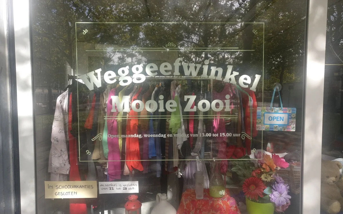 Etalage van kringloopwinkel met gekleurde kleding en speelgoed, winkelnaam zichtbaar op raam