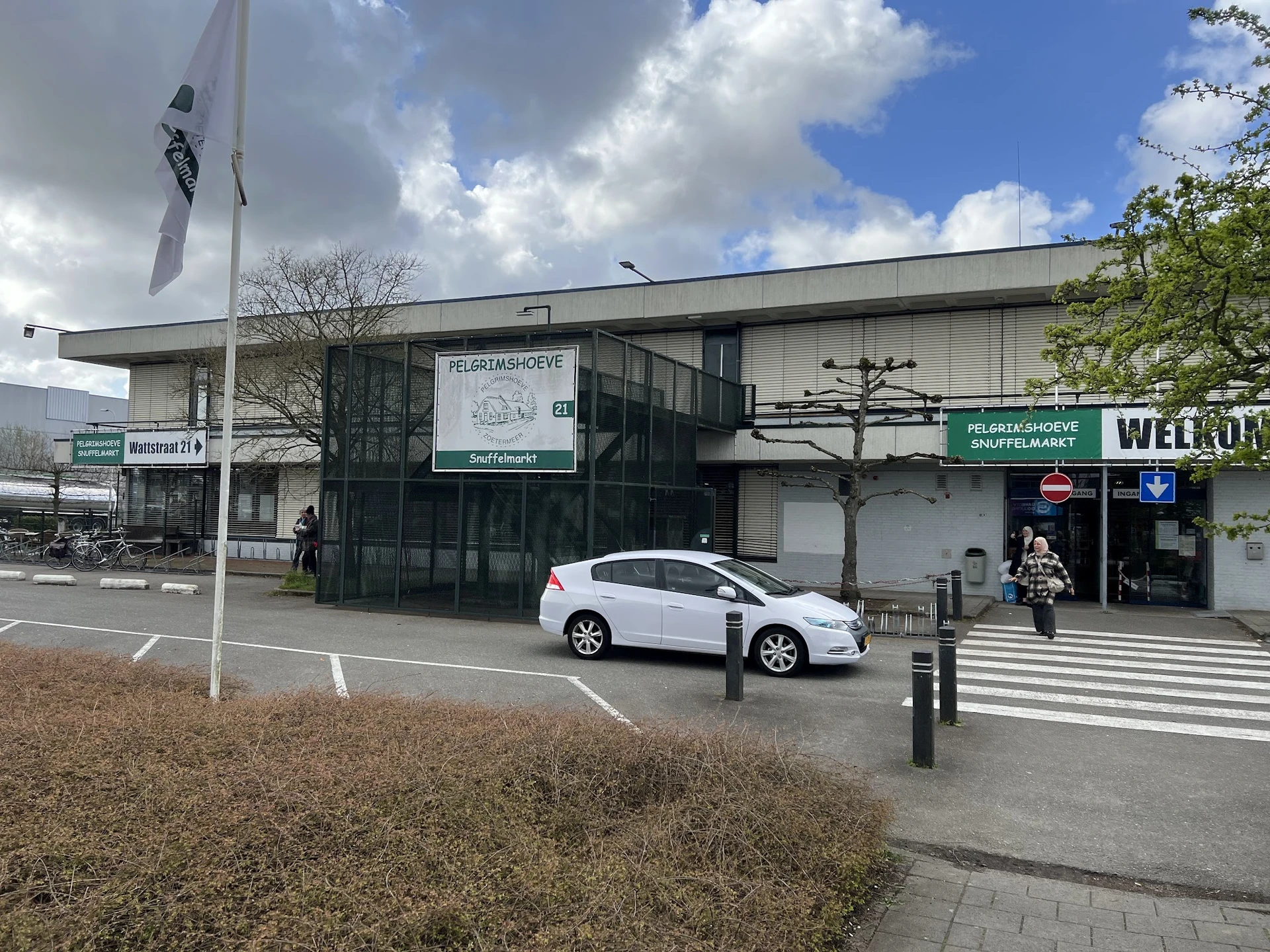 Gevel van kringloopwinkel Pelgrimshoeve met witte auto op parkeerplaats, automatische deuren zichtbaar