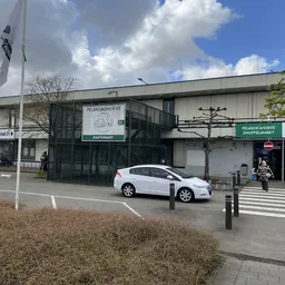 Gevel van kringloopwinkel Pelgrimshoeve met witte auto op parkeerplaats, automatische deuren zichtbaar