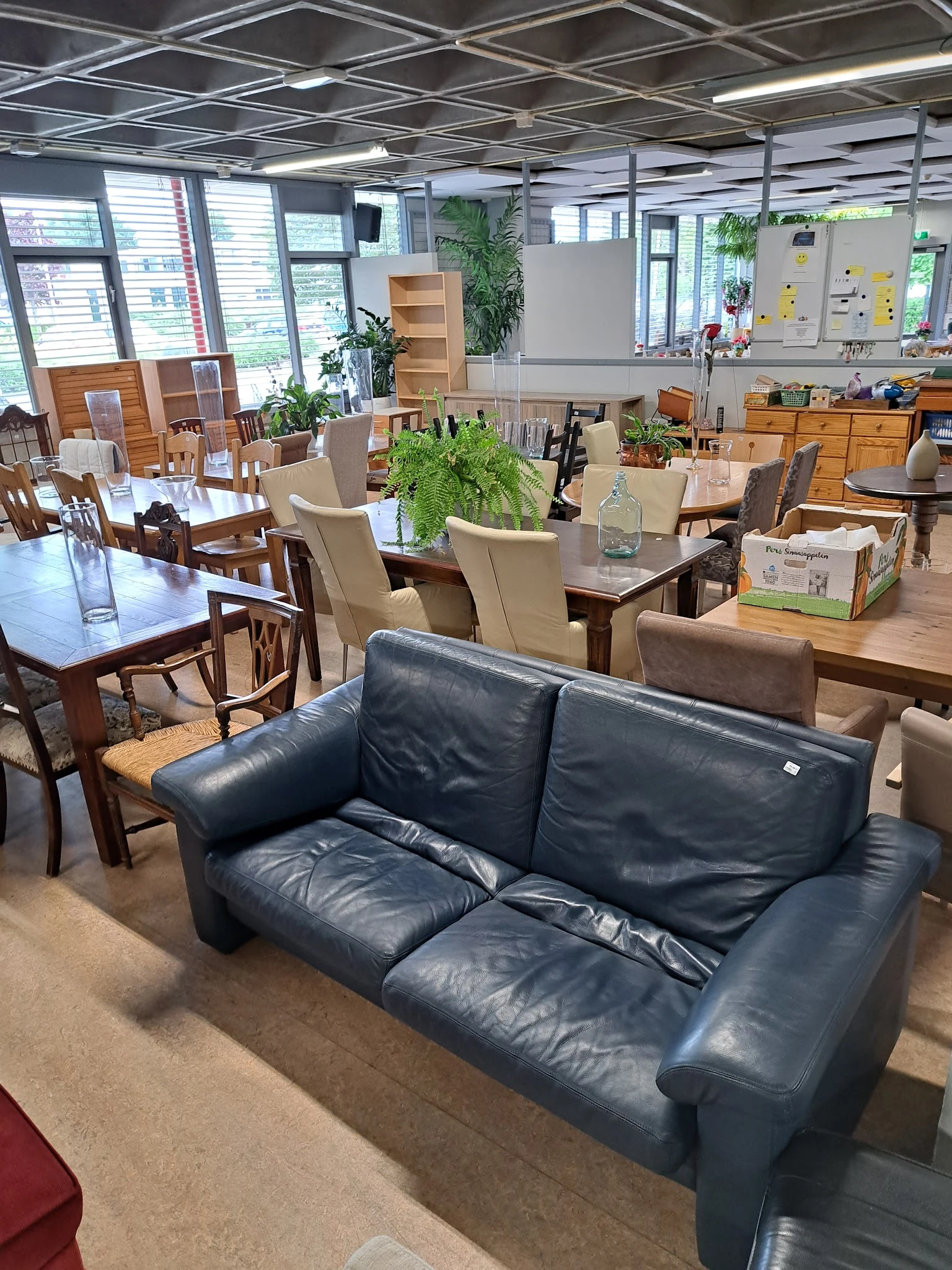 Winkelvloer met tweedehands meubels: een donkerblauwe leren bank op voorgrond, houten eettafels en stoelen, kast en planken m