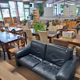 Winkelvloer met tweedehands meubels: een donkerblauwe leren bank op voorgrond, houten eettafels en stoelen, kast en planken m