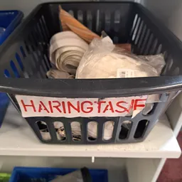 Zwarte plastic mand met wit serviesgoed en huishoudartikelen, gelabeld met rode tekst