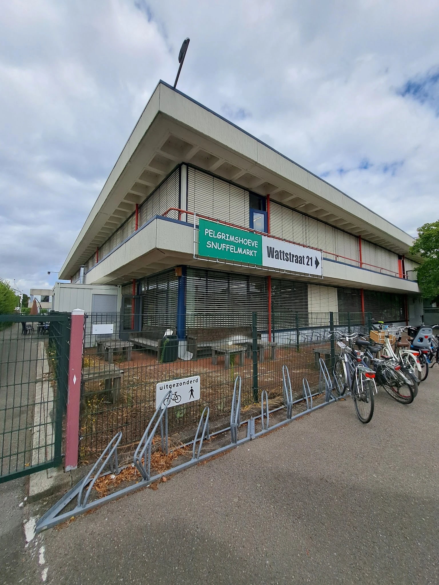 Voorgevel van kringloopwinkel Pelgrimshoeve met groene winkelnaambord, fietsenstalling met fietsen, en aangeven van Wattstraa