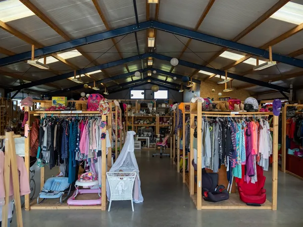 Overzicht van de winkelvloer met houten kledingrekken vol tweedehands kleding, babyartikelen en huishoudelijke spullen onder 