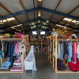 Overzicht van de winkelvloer met houten kledingrekken vol tweedehands kleding, babyartikelen en huishoudelijke spullen onder 