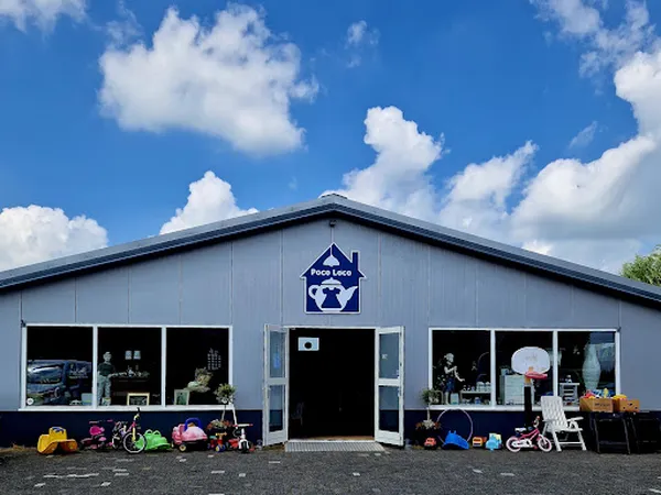 Grijze kringloopwinkel met winkelnaam op het dak, grote ramen, ingang in het midden, speelgoed en spullen buiten uitgestald