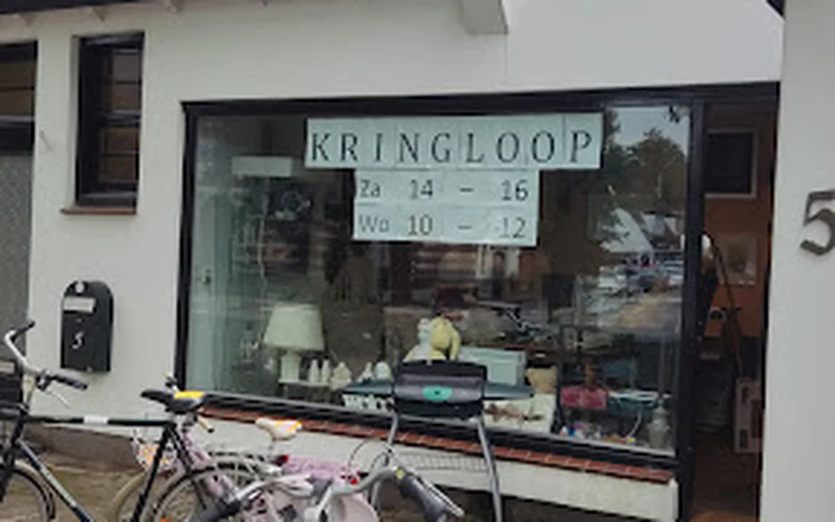 Witte gevel van kringloopwinkel met grote ramen, winkelnaam KRINGLOOP zichtbaar, openingstijden op raam, fiets ervoor